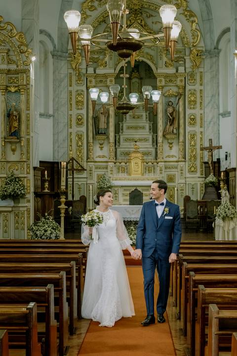Noivos a caminhar de mãos dadas no corredor central de uma igreja em Portugal, após a cerimónia de casamento. Decoração clássica, altar dourado e alegria do casal destacam o momento. casamento, igreja, noivos, cerimónia, fotografia de casamento.
'