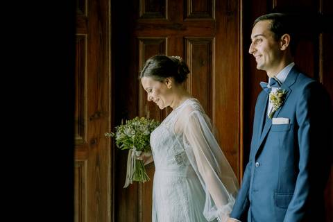 Noivos a saírem da cerimónia de casamento, com a noiva sorridente e bouquet de flores naturais, acompanhada pelo noivo elegante em fato azul. Fotografia de casamento romântica, destacando momentos de felicidade e o vestido de noiva bonito '