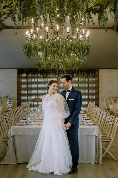 Noivos sorridentes num cenário elegante de casamento, com mesas decoradas, lustre imponente e detalhes em tons naturais. Ambiente ideal para celebrações únicas: fotografia de casamento, de casamento, fotógrafo profissional, decoração de casamento'