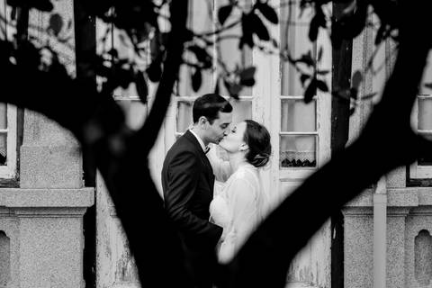 Noivos a beijarem-se num cenário romântico, enquadrados por um tronco de árvore, com fundo rústico de janelas antigas. Momento de amor captado com delicadeza.  fotografia de casamento, fotógrafo em Portugal, quinta de Santiago '