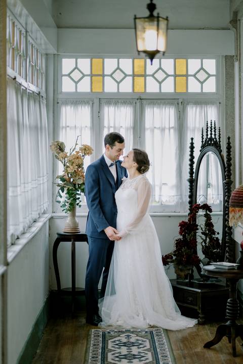 Noivos em momento íntimo e romântico numa varanda iluminada, rodeados por detalhes vintage e elegantes. A noiva com vestido rendado e o noivo de fato azul transmitem amor e cumplicidade. fotografia de casamento, fotógrafo em Portugal, quinta de Santiago'