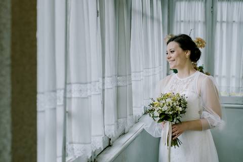 Noiva sorridente com vestido rendado e bouquet elegante, iluminada por luz natural junto a uma janela. Fotografia romântica e cheia de emoção. fotografia de casamento, noiva feliz, fotógrafo em Portugal, quinta de santiago'