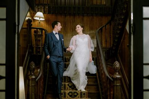 Noivos descem uma escadaria de madeira clássica, de mãos dadas, num momento de cumplicidade e alegria. A noiva brilha em vestido de renda delicado, enquanto o noivo exibe elegância no seu fato azul: fotografia de casamento, quinta de Santiago'