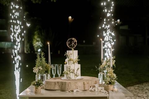 Imagem do bolo de casamento iluminado, rodeado de flores, velas e copos de champanhe, com destaque para os fogos de artifício. O cenário ao ar livre cria uma atmosfera mágica para este momento especial: bolo de casamento, decoração de casamento'