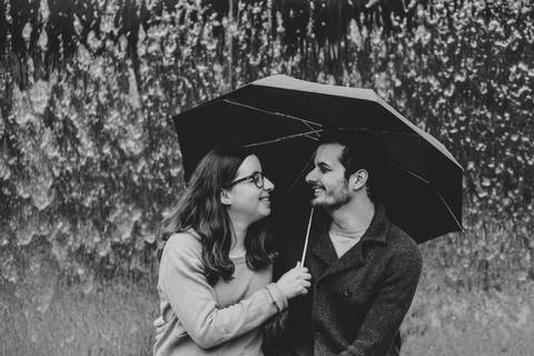 Casal sentado debaixo de um guarda-chuva preto, com fundo de cascata. Mulher segura o guarda-chuva e olha para o homem. fotografia de casamento, sessão de fotos de casamento, fotógrafo de casamentos, sessão em Bath, sessão em Bristol, sessão em Portishead'