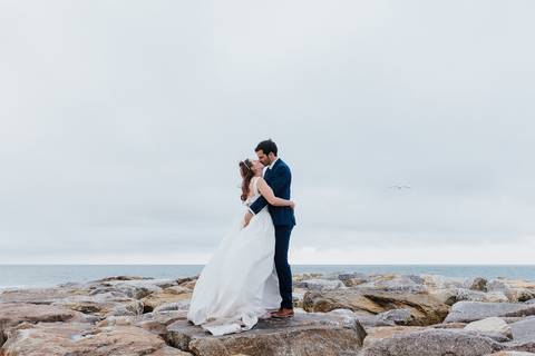 Fotografia de casamento na praia com noivos em pose romântica sobre rochas. Noiva com vestido branco e noivo com fato azul-marinho. Céu nublado e mar ao fundo. fotografia de pós casamento na praia de mira pelo fotógrafo de casamentos rui costa freire phot'