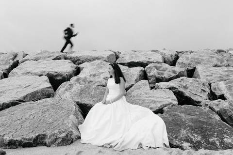 Fotografia de casamento na praia com noiva sentada em rochas e noivo a correr ao fundo. Imagem em preto e branco, capturando um momento único e espontâneo. Ideal para portfólio de fotógrafo de casamentos. fotografia de  pós casamento a preto e branco'