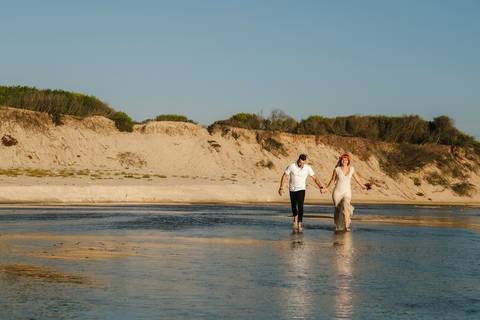 Casal de noivos a caminhar de mãos dadas na Praia da Tocha, atravessando águas rasas ao pôr do sol, com dunas naturais ao fundo. Sessão fotográfica de casamento na praia, captando momentos naturais: fotografia de casamento, fotógrafo de casamento.'