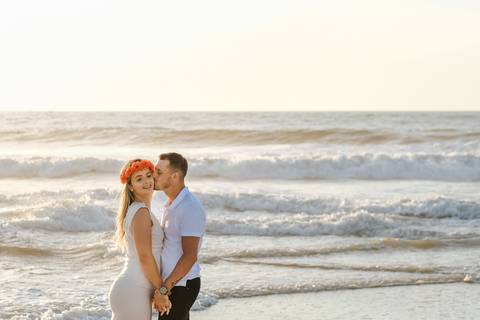Casal em sessão fotográfica na praia ao pôr do sol. Noiva com coroa de flores vermelhas e noivo em gesto carinhoso junto ao mar.  Palavras-chave: sessão de sorteiros, pré-casamento, sessão na praia, praia da tocha, rui costa freire Photography '