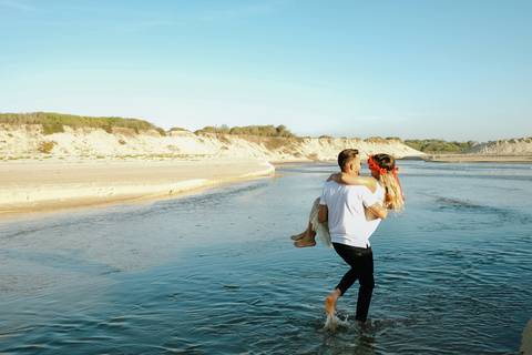 Noivo a carregar a noiva em momento romântico na Praia da Tocha, atravessando águas num cenário de dunas ao fundo. Fotografia de casamento na praia, destacando a naturalidade. sessão fotográfica, Praia da Tocha, fotografia de casamento, rui costa freire'