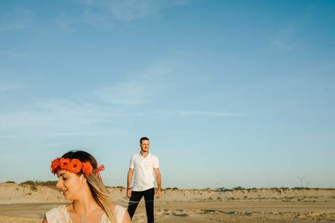 Casal em sessão fotográfica ao pôr do sol numa praia deserta, noiva com coroa de flores vermelhas e o noivo casual ao fundo. Cenário natural perfeito para fotos de pré-casamento. Praia da Tocha, pré-casamento, sessão fotográfica, fotos na praia.'