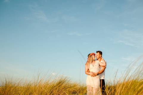 Namorados em sessão de solteiros ao pôr do sol, entre as dunas. Ela usa um vestido rendado e coroa de flores, ele abraça-a com ternura. Ambiente natural,  fotografia de casamento, pré-casamento, sessão pré Casamento.'