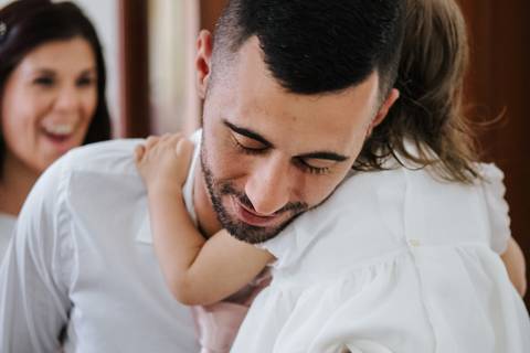 no dia do baptismo o pai abraça a criança e a mãe olha e sorri'