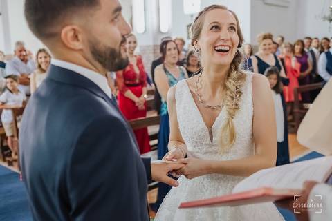 Noiva coloca a aliança no dedo do noivo e sorri.'