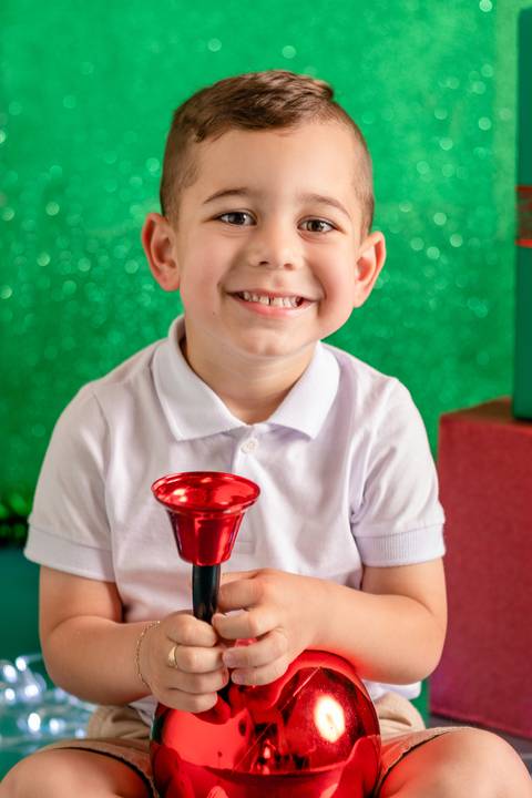 Cenário de natal decorado com presentes, com menino de 3 anos sentado e sorrindo, com sino e bola de natal'