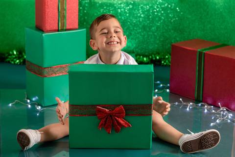 Cenário de natal verde decorado com presentes, com menino de 3 anos sentado e sorrindo, atrás de um presente de natal'