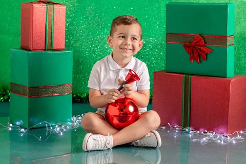 Cenário de natal verde decorado com presentes, com menino de 3 anos sentado e sorrindo, com sino e bola de natal'