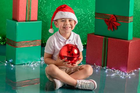 Cenário de natal verde decorado com presentes, com menino de 3 anos sentado e sorrindo, com touca de papai noel e bola de natal'