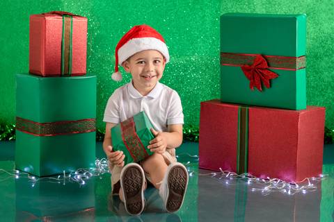 Cenário de natal verde decorado com presentes, com menino de 3 anos sentado e sorrindo, com touca de papai noel e segurando um presente'
