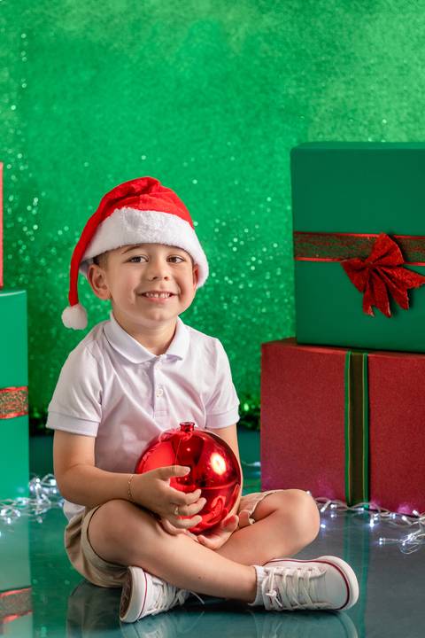 Cenário de natal verde decorado com presentes, com menino lindo de 3 anos sentado e sorrindo, com touca de papai noel e bola de natal'