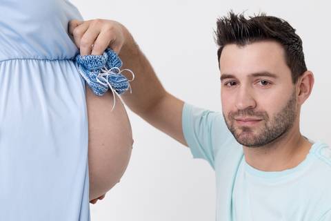 Casal gravidos de 9 meses com o papai segurando o sapatinho do bebê com a mamãe de vestido azul em ensaio em estúdio com o fundo branco ensaio fotógrafo Ander Lopes em Taquara/RS'