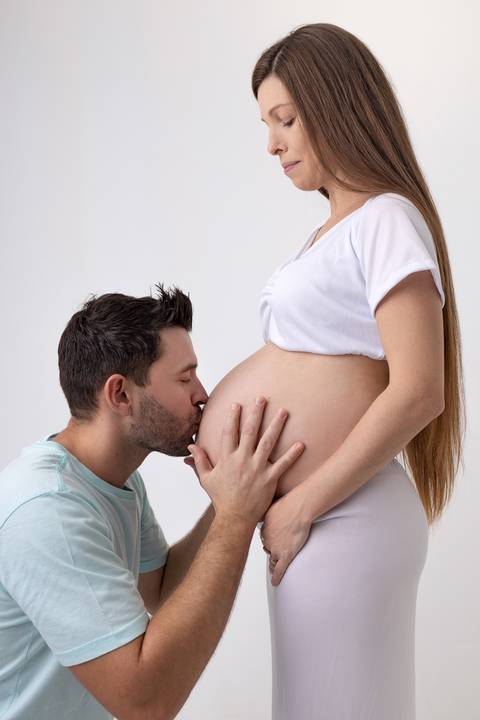 Casal gravidos de 9 meses com o papai beijando a barriga e com a mamãe de vestido branco em ensaio em estúdio com o fundo branco ensaio fotógrafo Ander Lopes em Taquara/RS'