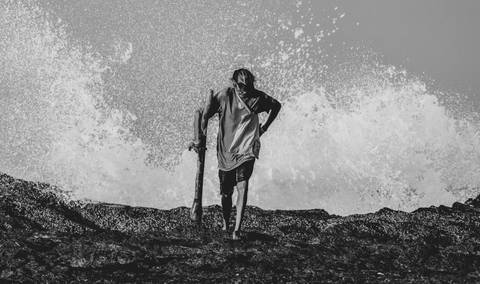 homem segundando um didgereedoo andando em pedras com ondas batendo atrás'