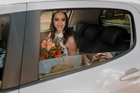 A noiva chega ao local do casamento sorrindo, carregando o buquê em mãos e demonstrando a felicidade de viver esse dia especial.'