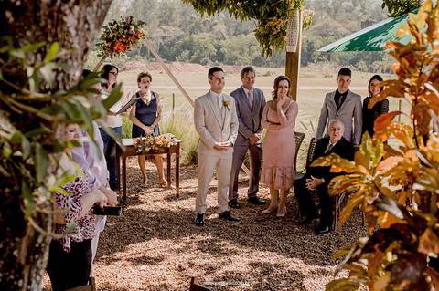 O noivo, cercado por familiares e amigos, aguarda ansioso a entrada da noiva na cerimônia intimista ao ar livre.'