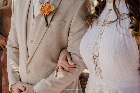 Detalhe das mãos da noiva entrelaçadas ao braço do noivo, simbolizando união e compromisso.'