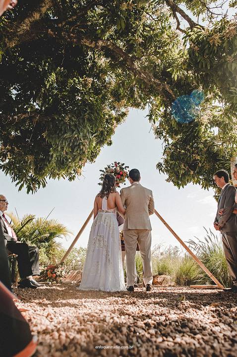 Aos pés de uma grande árvore, os noivos escutam as palavras do celebrante, em um cenário natural e acolhedor.'