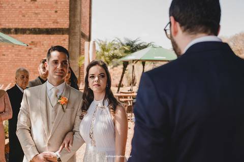 Juntos, de mãos dadas, os noivos ouvem a leitura do irmão da noiva atentos, com serenidade e emoção no olhar.'