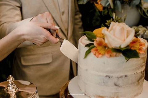 As mãos dos noivos se unem no corte do bolo de casamento, simbolizando parceria e união em um momento cheio de significado.'
