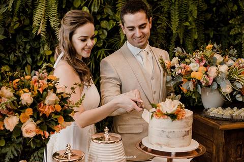 Gláucia e Fernando, sorridentes, compartilham a tradição do corte do bolo em meio à decoração florida do casamento intimista no Villa Itália, Piracicaba.'