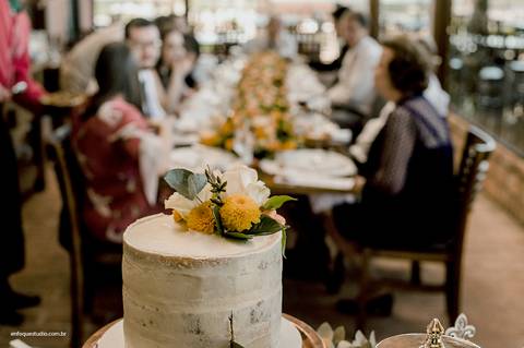 O bolo com flores naturais enfeita a mesa da recepção, enquanto familiares e convidados aguardam o brinde.'