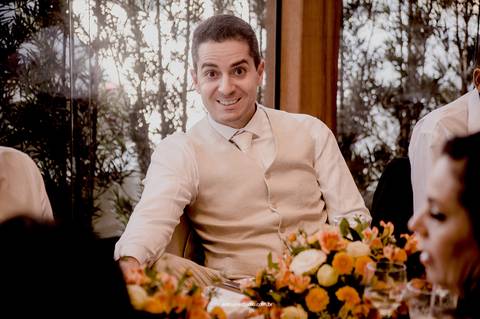 O noivo se diverte entre conversas à mesa, registrando a leveza e a alegria do almoço de casamento.'