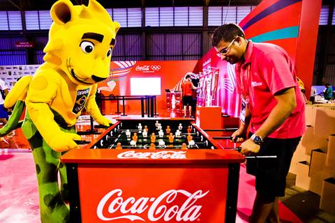 	Fotografia do mascote olímpico Ginga brincando em uma mesa de pebolim da Coca Cola, patrocinadora oficial dos Jogos Escolares da Juventude em Londrina.'