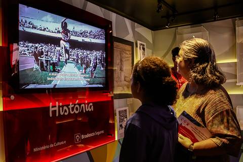 	Fotografia Institucional de uma senhora e uma criança assistem um vídeo de atletismo em televisão instalada no Museu Itinerante do Bradesco Seguros.'