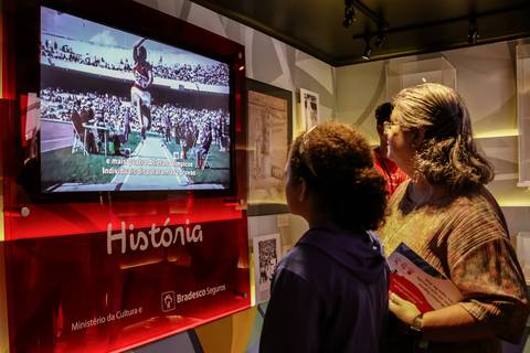 A fotografia corporativa mostra uma mulher e menina que assistem vídeo de uma prova de salto à distância, a peça faz parte da exposição do Museu Itinerante que está em Londrina e patrocinado pelo Bradesco Seguros e Ministério da Cultura.'