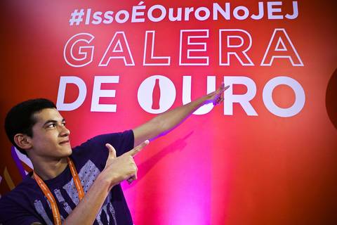 Jovem atleta imita gesto do velocista Usain Bolt em frente a um painel e publicidade da Coca Cola, fotografia institucional publicitária dos Jogos da Juventude.'