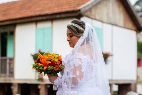 Noiva com Vestido em renda com um buquê laranja nas mãos em frente a uma casa rústica na Rota do Lagarto'