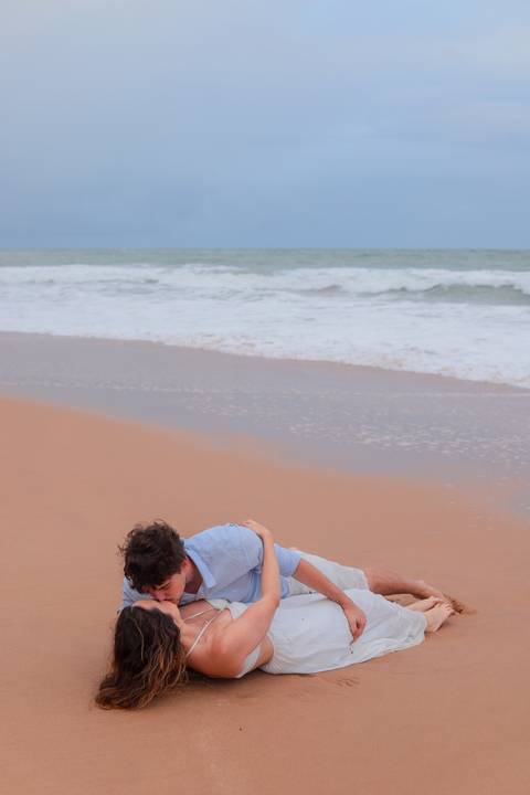 Noivos rolando na areia, rindo e vivendo o momento. Um ensaio que mostra que o verdadeiro amor também se diverte.'