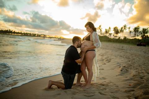 Toque simbólico na barriga da gestante com pôr do sol ao fundo, no deck de madeira em Barra do Jacuípe, fotografado por Waldyr Lantyer, fotógrafo de famílias na região da Praia do Forte Bahia e Iberostar.'