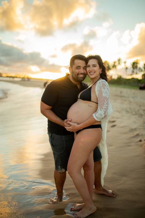 Toque simbólico na barriga da gestante com pôr do sol ao fundo, no deck de madeira em Barra do Jacuípe, fotografado por Waldyr Lantyer, fotógrafo de famílias na região da Praia do Forte Bahia e Iberostar.'