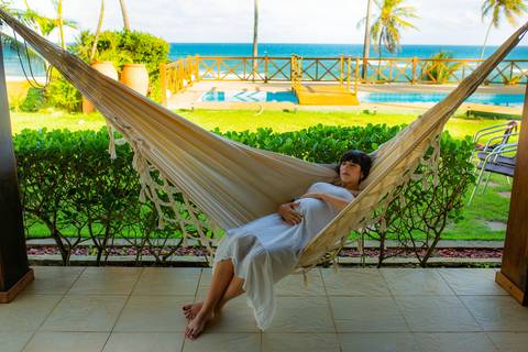 Gestante de branco deitada na rede com o marido em casa de praia em Barra do Jacuípe, Bahia — ensaio ao ar livre à espera do primeiro filho. '