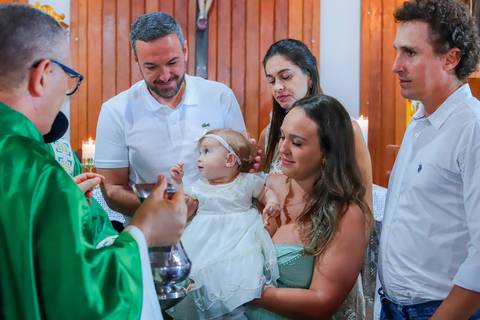 Pais emocionados segurando a bebê Catarina durante o batizado em Praia do Forte, com cenário encantador e luz suave da manhã.'