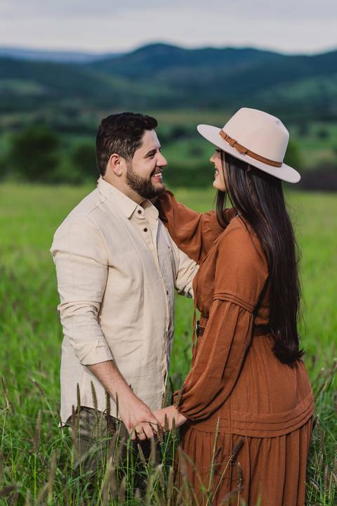 Jaque e Mateus durante ensaio pré-casamento em área rural, com campo verde e montanhas ao fundo.'