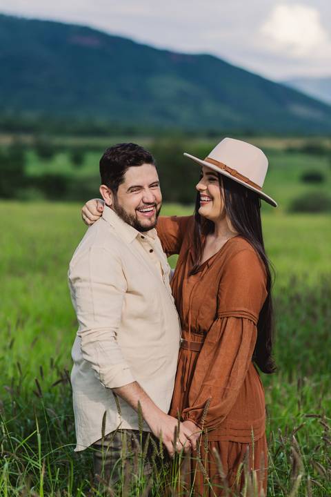 Jaque e Mateus sorrindo e abraçados em meio à paisagem verde da zona rural de Urandi, Bahia.'