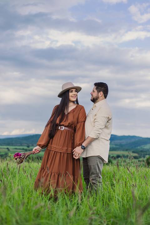 Jaque e Mateus caminhando juntos pelo campo durante o pré-casamento, com serra e céu azul ao fundo.'