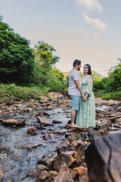Jaque e Mateus juntos em um riacho cercado por pedras e vegetação durante o ensaio pré-casamento.'